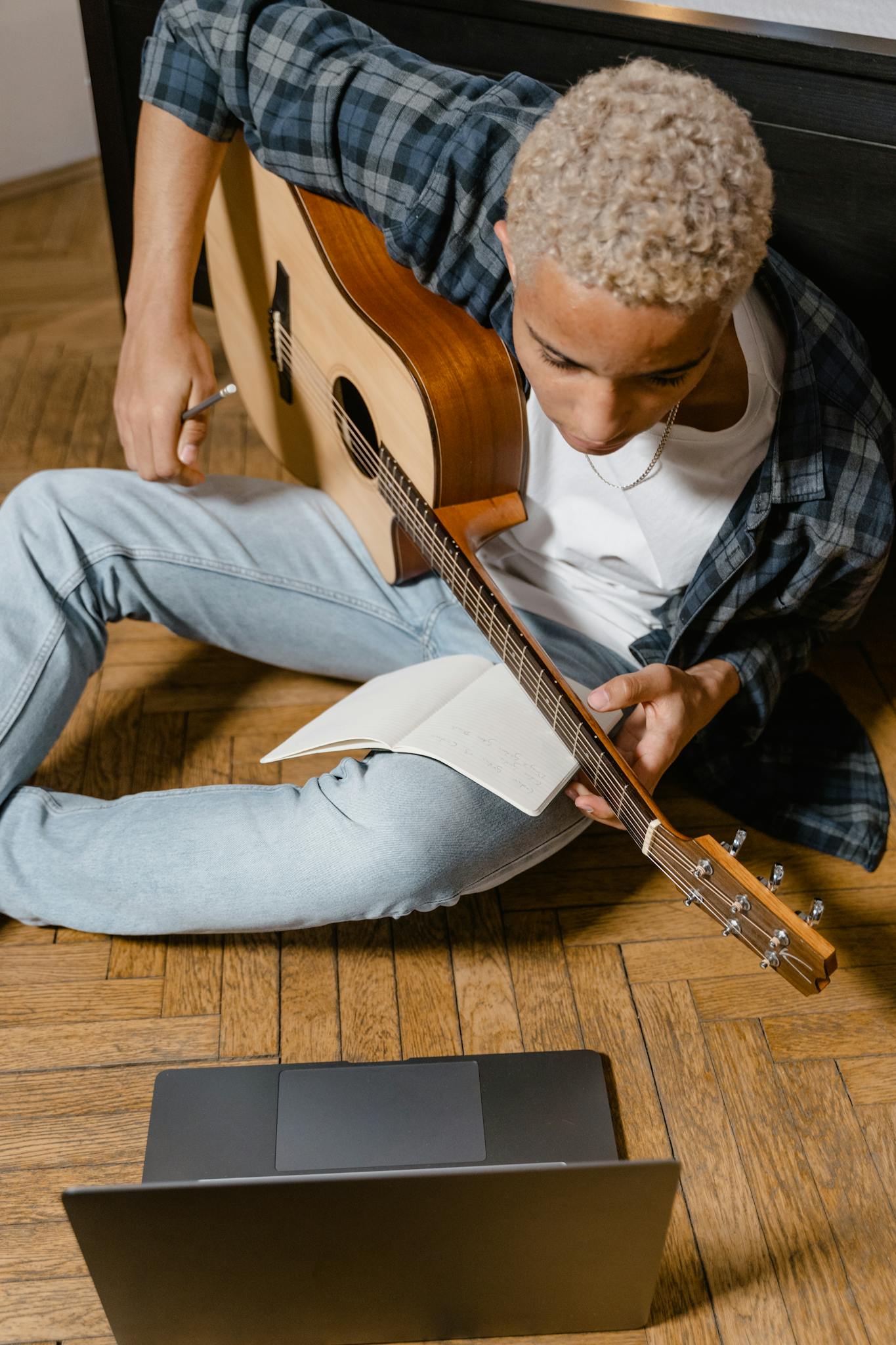 A young man learning guitar online, sitting on the floor with a laptop and notebook.
