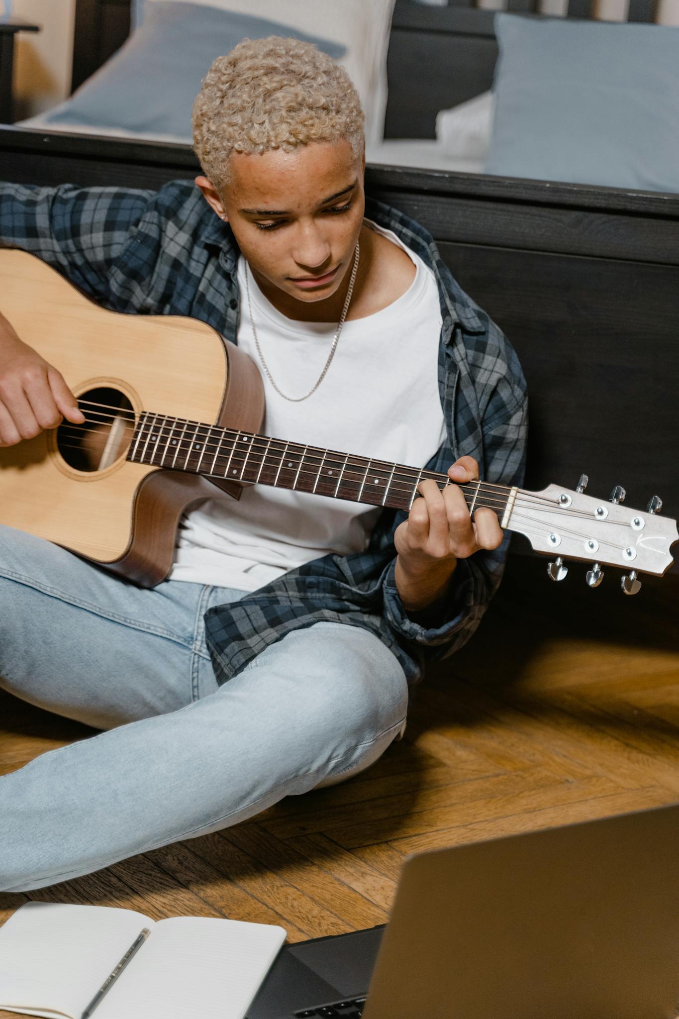 A young man practicing guitar at home while watching an online lesson on a laptop.