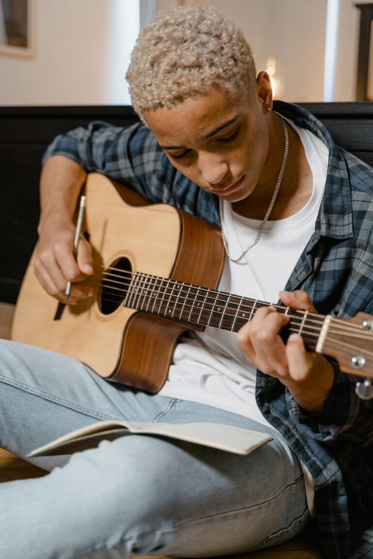 A young man sits in a cozy room playing an acoustic guitar while learning from a notebook.