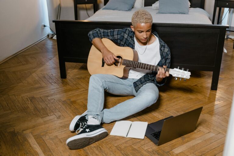 Man sits on bedroom floor playing guitar while engaging in online music class.
